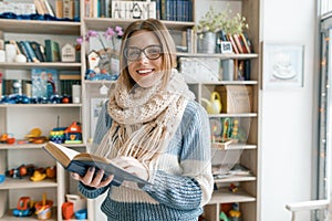 Winter autumn portrait of young beautiful girl student wearing glasses in knitted warm scarf and sweater reading book indoors