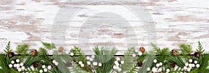 Winter bottom border with evergreen branches, white berries and pine cones on a rustic white wood banner background photo