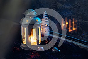 Winter Christmas Eve. Frosted window with Christmas decoration. Lantern with a lit candle near the window with frosty patterns on