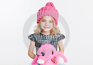 Winter clothes. Portrait of little curly girl in knitted pink winter hat isolated on white. Pink rabbit toy in her hands