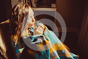 Winter cold concept. Young freezing woman in comfortable chair warming frozen hands wrapped in warm fluffy woollen plaid blanket.