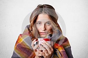 Winter cold sickness concept. Freezing beautiful woman wrapped in warm checkered plaid blanket, drinks hot beverage, tries to warm