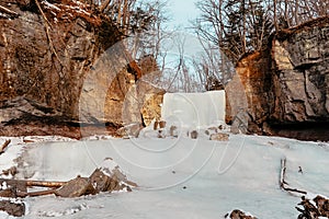 Winter cold tourist landscape, a frozen waterfall among mountains, around a forest without leaves and a lot of white snow