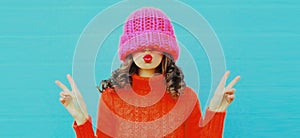 Winter colorful portrait beautiful young woman blowing her lips wearing red knitted sweater, pink hat on blue background