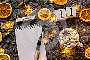 Winter composition. Wooden calendar January 11th Cup of cocoa with marshmallow, empty open notepad with pen, dried oranges, light