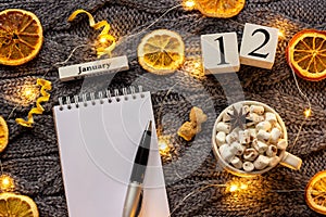 Winter composition. Wooden calendar January 12th Cup of cocoa with marshmallow, empty open notepad with pen, dried oranges, light