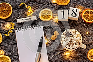 Winter composition. Wooden calendar January 18th Cup of cocoa with marshmallow, empty open notepad with pen, dried oranges, light