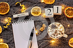 Winter composition. Wooden calendar January 21st Cup of cocoa with marshmallow, empty open notepad with pen, dried oranges, light