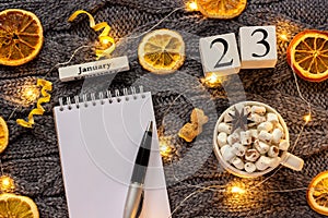 Winter composition. Wooden calendar January 23rd Cup of cocoa with marshmallow, empty open notepad with pen, dried oranges, light