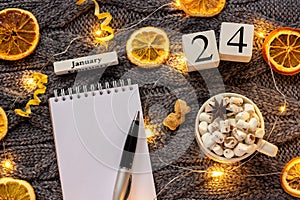 Winter composition. Wooden calendar January 24th Cup of cocoa with marshmallow, empty open notepad with pen, dried oranges, light