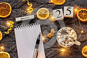 Winter composition. Wooden calendar January 25th Cup of cocoa with marshmallow, empty open notepad with pen, dried oranges, light