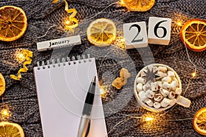 Winter composition. Wooden calendar January 26th Cup of cocoa with marshmallow, empty open notepad with pen, dried oranges, light