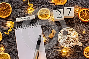 Winter composition. Wooden calendar January 27th Cup of cocoa with marshmallow, empty open notepad with pen, dried oranges, light
