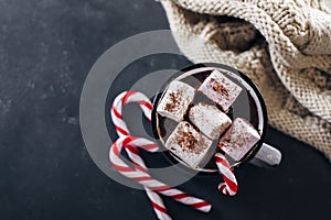 Winter cozy beverage. Glass with hot chocolate with marshmallow, candy cane, cozy warm scarf on dark blue background. Top view