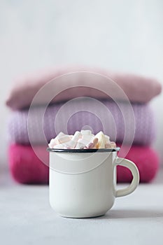 Winter cozy concept. Sweater weather. Coffee with marshmallows in white enameled metal cup on the background of stack of pink