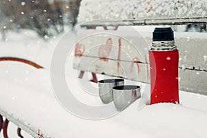 Winter Date Ideas for Couples. Winter holidays, hot drinks concept. Red thermos and cups on a snowy bench in winter park