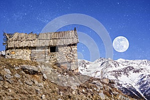 Winter evening landscape in french Alps. Old stone abandoned house on the mountain. Full moon on the sky, starry background