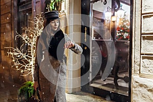 Winter fashion. Christmas, New Year concept. Woman waiting friends on city street by holiday decorated cafe showcases.