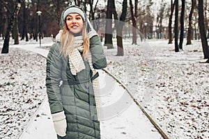 Winter fashion. Young woman wearing long green coat with scarf, hat, mittens in snowy park.