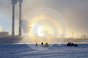 Winter fishing, men`s passion, fishermen catch fish on a frozen river against the background of the factory pipes at sunset.