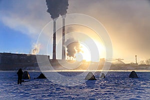 Winter fishing, men`s passion, fishermen catch fish on a frozen river against the background of the factory pipes at sunset.