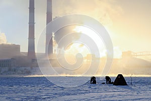 Winter fishing, men`s passion, fishermen catch fish on a frozen river against the background of the factory pipes at sunset.