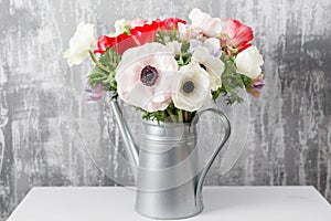 Winter flowers. Anemones in a vase watering can standing on a wooden table. On the background old gray wall art. copy