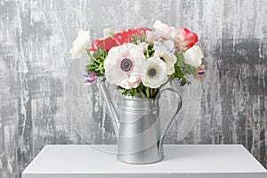 Winter flowers. Anemones in a vase watering can standing on a wooden table. On the background old gray wall art. copy