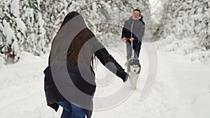 In the winter forest, the girl stands with her back with her hands open, and a man with a husky dog runs to meet her. A