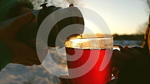 In winter forest man pours hot tea from thermos into glass goblet at sunset bright sun. girl holding glass of tea in