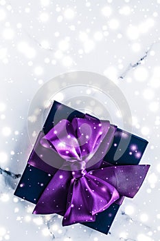 Winter holiday gift box with purple silk bow, snow glitter on marble background as Christmas and New Years presents for luxury