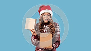 Winter holiday surprise. Pretty millennial woman in Christmas outfit opening gift box on blue studio background