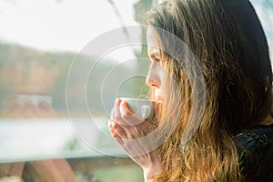 Winter holidays, christmas, hot drinks and people concept. Happy beautiful young woman drinking coffee behind a window. Horizontal