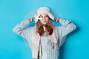 Winter and holidays concept. Happy redhead girl in beanie hat, scarf and gloves showing peace sign and pucker lips sassy