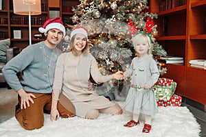 Winter holidays with family. Smiling Caucasian mother and father in Santa hats with baby girl by decorated Christmas tree at home