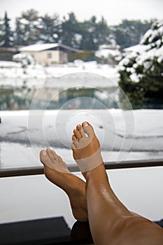 Winter holidays, resting pretty woman bare feet and legs lying up on stylish table, window view of snow landscape and frozen lake
