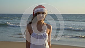 Winter holidays in a warm country. Young adult woman in Santa Claust Hat stands on the beach. Portrait of a long-haired