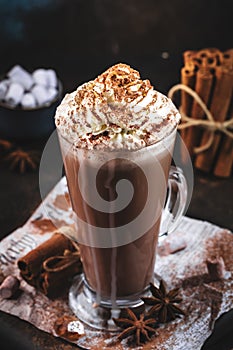 Winter hot chocolate cocoa drink in tall glass with ispices and whipped cream on black table. Dark blue background. Copy space