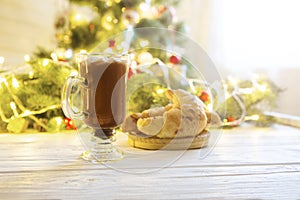 Winter hot chocolate with marshmallows and a croissant on a white wooden table against the background of the Christmas tree