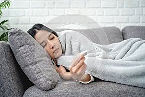 Winter Illnesses. Young sick Asian woman taking temperature with thermometer while lying under blanket on sofa at home