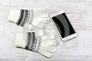 Winter knitted gloves and smart phone with blank screen on white wooden background. Smartphone mock-up with blank black screen