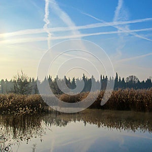 Winter lake landscape in early winter morning with blue sky and plane tracks reflected in water