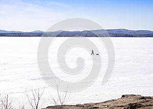 Winter landscape, a fisher goes by frozen lake with his staff, mountains on a skyline. Blue sky, sunny cloudless day.