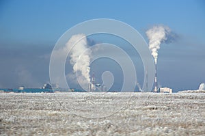 Winter landscape with metallurgical plant with heavy smoke from pipes behind a field covered with frozen dry grass under blue sky