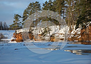 Winter landscape with red sandstone cliffs on the bank of the river Salaca, the sun shines on the trees and the river bank, the