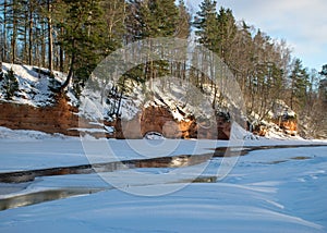 Winter landscape with red sandstone cliffs on the bank of the river Salaca, the sun shines on the trees and the river bank, the