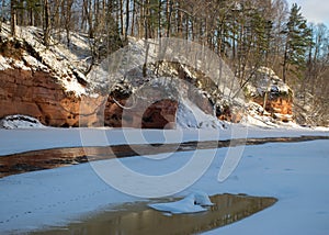 Winter landscape with red sandstone cliffs on the bank of the river Salaca, the sun shines on the trees and the river bank, the