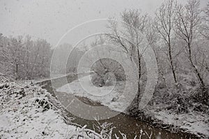 Winter landscape with snow coming down and a river in the middle of trees covered by snow