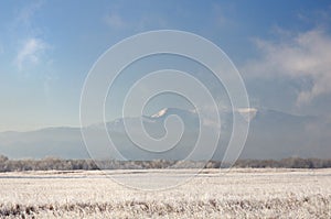 Winter landscape with snowy mountains behind field covered with frozen dry grass under dark blue sky during sunrise