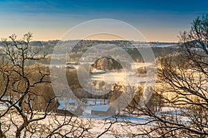 Winter landscape of snowy village near misty river at sunrise time